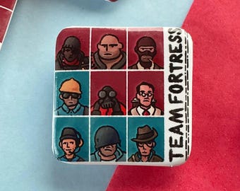 May include: Square Team Fortress pins with a grid of character portraits in red and teal. The pins have the words "TEAM FORTRESS" printed on the side. The background is a mix of red and blue.