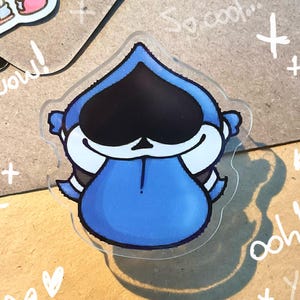 Può includere: Un adesivo trasparente in acrilico con un personaggio dei cartoni animati blu con un viso nero e un copricapo a forma di corona. Scarabocchi bianchi, tra cui cuori, croci e le parole "wow!", "ooh!" e "Yay!", circondano l'adesivo.