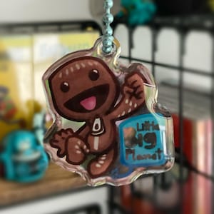 May include: A clear acrylic charm featuring a brown cartoon character with a smiling face and the text "Little Big Planet." The charm is hanging from a light blue beaded chain.