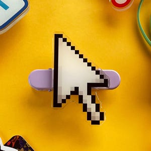 May include: A pixelated white and black computer cursor pin with a light purple backing, set against a bright yellow background. Other colourful office supplies, including a pink binder clip and rubber bands, are scattered around the pin.