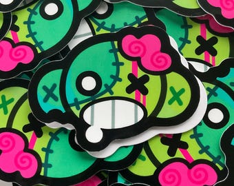 May include: A collection of stickers featuring a cartoon zombie head design. The stickers have a green base with black outlines, pink brain details, and white accents. The design includes cross-shaped stitches and a stitched-up appearance.