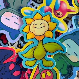 May include: A collection of colorful cartoon stickers featuring a smiling yellow sunflower with green leaves, a blue character with a white face, a red and white character with a yellow eye, and a green character with a white face.