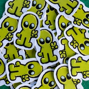 May include: A pile of green alien stickers with black outlines and white eyes. The aliens are all making a peace sign with their hands.