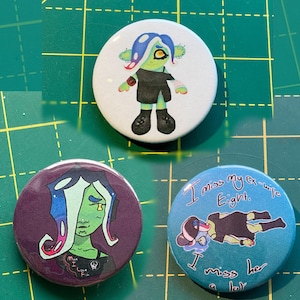 May include: Three circular badges with cartoon character illustrations. One shows a green character with blue hair and a black outfit. Another has a purple background with a green character. The third badge has the text "I miss my ex-wife Eight."