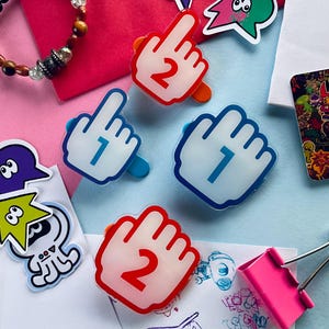 Puede incluir: Una colección de artículos de papelería coloridos, que incluyen clips de papel en forma de mano con los números 1 y 2, y varias pegatinas de dibujos animados. También se ve un clip rosa y una pulsera de cuentas. El fondo es una mezcla de rosa y azul.