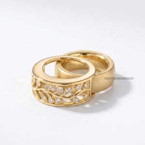 May include: Two gold rings, one featuring an intricate openwork leaf pattern set with small round diamonds, and the other a plain, smooth gold band. The text NEXTDIAMONDSCO is visible on the side of the plain ring, showcasing a polished finish.