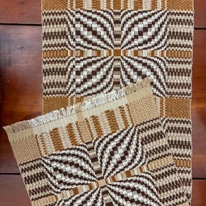 May include: Two rectangular woven table runners featuring a geometric pattern in shades of brown, tan, and cream. The design includes a central checkered motif with wavy lines and fringed edges on the top and bottom of each textile piece.
