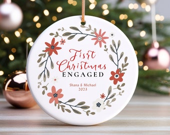 May include: White ceramic Christmas bauble featuring a red floral wreath and the text "First Christmas Engaged" with the names Shana & Michael and the year 2023.