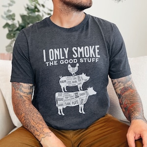May include: A dark heather grey t-shirt with a white graphic of a pig, chicken, and cow with labelled cuts of meat. The text "I Only Smoke The Good Stuff" is printed on the shirt.