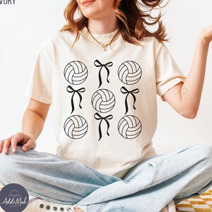 May include: A white t-shirt with a black and white design of volleyballs with bows. The shirt is ivory in colour.