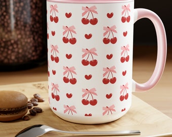 May include: A white ceramic mug with a pink rim and handle. The mug features a repeating pattern of red cherries with pink bows and hearts.