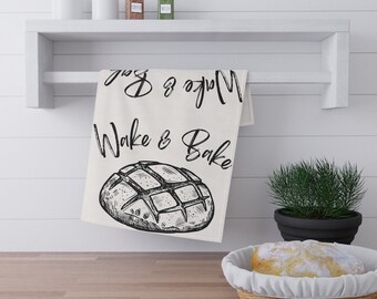 Op de afbeelding: Een crèmekleurige keukendoek met de woorden "Wake & Bake" in een scriptlettertype, samen met een schets van een brood. De handdoek hangt aan een witte plank boven een houten aanrecht. Andere items zijn potten en een plant.