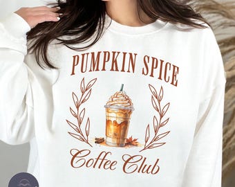 May include: White sweatshirt with the text "PUMPKIN SPICE Coffee Club" in brown. The design features a coffee drink with whipped cream, surrounded by a wreath of leaves. A fall-themed graphic tee.
