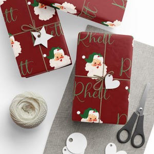 May include: Red wrapping paper with a repeating pattern of vintage Santa Claus faces and the name "Rhett" written in green cursive.