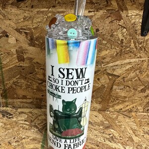 May include: A white tumbler with a colorful design featuring sewing supplies and a black cat. The text on the tumbler reads "I sew so I don't choke people. Save a life, send fabric."