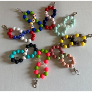 May include: A collection of beaded wristlet keychains in various colors. Designs include red, white, and blue, baseball-themed, and solid colors like turquoise, black, and pink. Each keychain has a silver-colored clasp.