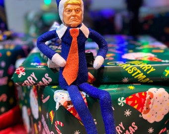 Może przedstawiać: Niebieska lalka elfa z żółtą twarzą i pomarańczowym krawatem siedzi na zielonym pudełku prezentowym z wzorem Merry Christmas. Pudełko jest zapakowane w zielony papier z białym wzorem śnieżynek.