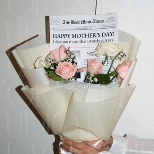 以下が含まれることがあります： クリーム色の紙に包まれたピンクと白のバラの花束。「HAPPY MOTHER'S DAY! I love you more than words can say」というテキストが入った新聞スタイルのカードが含まれています。花束はピンクのリボンで結ばれています。