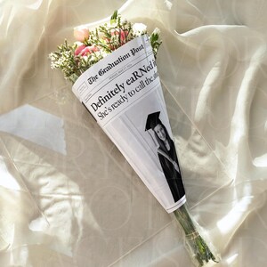 May include: A bouquet of flowers wrapped in a newspaper cone. The newspaper reads "The Graduation Post" with the headline "Definitely earNed" and a photo of a graduate. The flowers include pink and white blooms with green foliage.