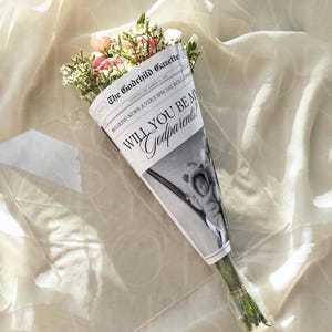 Puede incluir: Un ramo de flores rosas y blancas envuelto en un cono de periódico. El periódico dice "The Godchild Gazette" y la pregunta "Will You Be My Godparents?" sobre un fondo de tela crema.