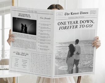 May include: A black and white newspaper titled "The Lover Times" with the headline "ONE YEAR DOWN, FOREVER TO GO!" The newspaper features a photo of a couple on a beach and a word search puzzle. The newspaper is being held up.