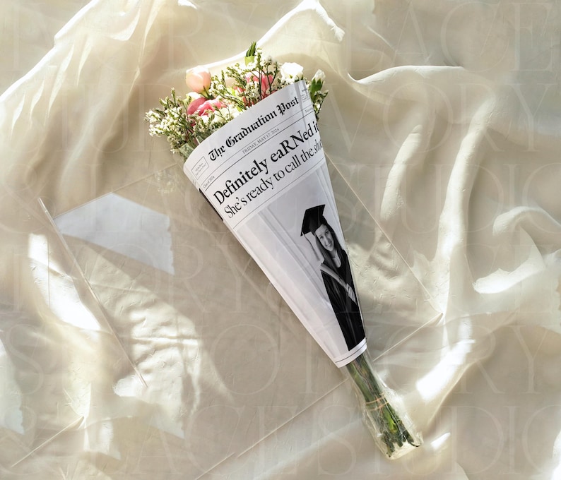 May include: A bouquet of flowers wrapped in a newspaper cone. The newspaper reads "The Graduation Post" with the headline "Definitely earNed" and a photo of a graduate. The flowers include pink and white blooms with green foliage.