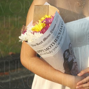 May include: A bouquet of colorful flowers wrapped in newspaper. The newspaper reads "Happy Mother's Day!" and features a photo. The bouquet includes white, yellow, and pink flowers.