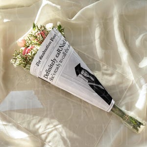 May include: A bouquet of pink and white flowers wrapped in newspaper. The newspaper reads "The Graduation Post" with the headline "Definitely eARNed it." A photo of a graduate is also visible.