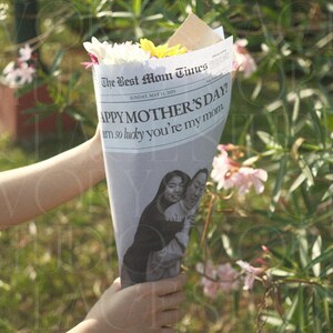 May include: A bouquet of white and yellow flowers wrapped in a newspaper cone. The newspaper reads "HAPPY MOTHER'S DAY!" with a photo of two women. The bouquet is held in front of a green, leafy background.