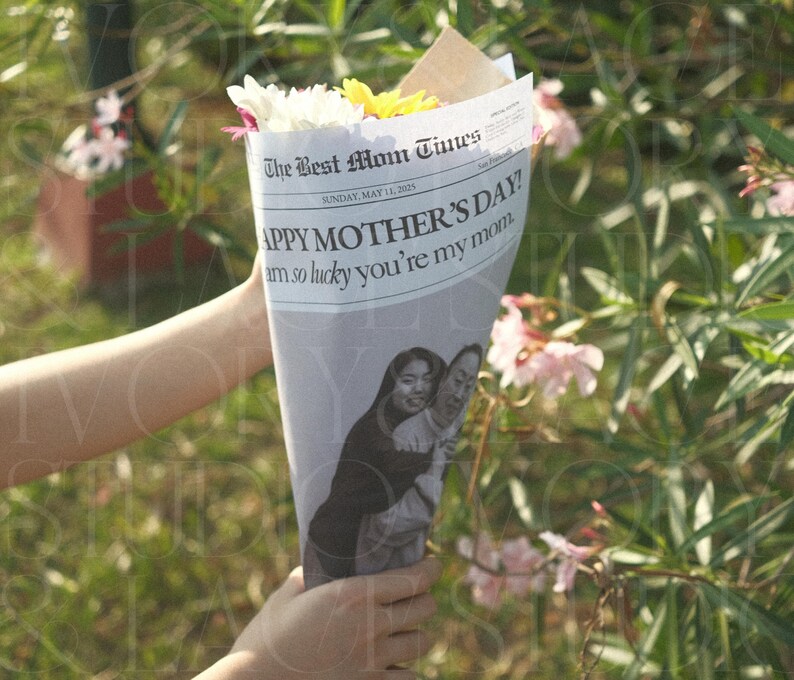 May include: A bouquet of white and yellow flowers wrapped in a newspaper cone. The newspaper reads "HAPPY MOTHER'S DAY!" with a photo of two women. The bouquet is held in front of a green, leafy background.