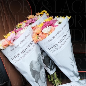 May include: Bouquets of colorful flowers, including pink, yellow, and orange blooms, are wrapped in newspaper-style paper. The paper reads "HAPPY MOTHER'S" and "The Best Mom Times". The bouquets are a thoughtful gift.