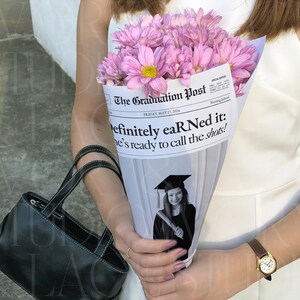 May include: A bouquet of pink flowers with yellow centers wrapped in a newspaper cone. The newspaper reads "The Graduation Post" with the headline "Definitely eaR Ned it: he's ready to call the shots!" A black handbag is in the foreground.