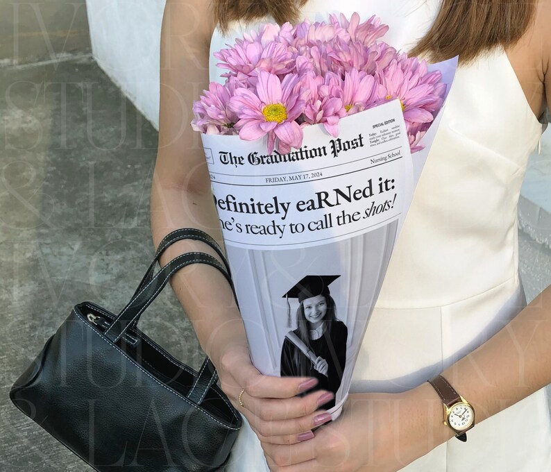 May include: A bouquet of pink flowers with yellow centers wrapped in a newspaper cone. The newspaper reads "The Graduation Post" with the headline "Definitely eaR Ned it: he's ready to call the shots!" A black handbag is in the foreground.