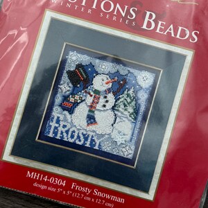 May include: A cross-stitch kit with a snowman design. The kit, titled "Frosty Snowman," features a framed image of a snowman wearing a scarf and hat. The design size is 12.7 cm x 12.7 cm. The packaging is red with the words "Buttons & Beads".
