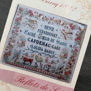 Op de afbeelding: Een gedrukt patroon voor kruissteekborduurwerk, met de tekst "Louisa Barny 1892" en "Reflets de Soie". Het ontwerp bevat een centraal geborduurd beeld met vogels, bloemen en tekst in het Frans. Het kleurenpalet omvat beige, rood en groen.