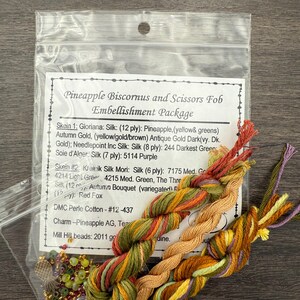 May include: A Pineapple Biscornus and Scissors Fob Embellishment Package containing three twisted skeins of embroidery thread in variegated shades of green, gold, brown, and purple, along with a small bag of assorted beads and a metal pineapple charm.