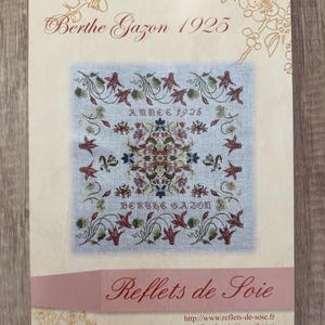 May include: A cream-colored paper with a floral design border. The center features a cross-stitch pattern with a floral design in shades of red, green, and blue. The text "Berthe Gazon 1925" is at the top of the paper. The text "Reflets de Soie" is at the bottom of the paper. The text "http://www.reflets-de-soie.fr" and "refletsdesoie@gmail.com" are below the text "Reflets de Soie".