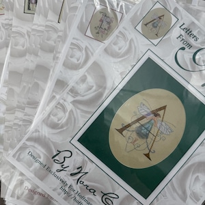 May include: A stack of cross-stitch patterns in clear plastic sleeves. The designs feature an embroidered letter "A" with a fairy, and the text "Letters From" and "By Nora Corbett". The background is a white rose pattern.