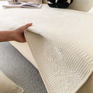 May include: Close-up of a white quilted sofa cushion with a floral pattern border. The cushion is being lifted to show the underside.