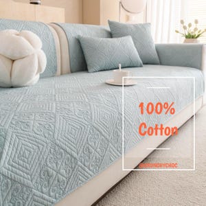 May include: A pale blue quilted sofa cover with matching cushions. The cover has a geometric design and the text "100% Cotton". A white decorative pillow and a teacup and saucer are also visible.
