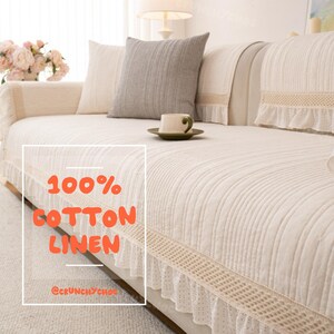 May include: A cream-colored sofa cover featuring a vertical ribbed texture, a decorative crocheted trim, and a ruffled lace edge. The fabric is labeled with the text 100% COTTON LINEN and @CRUNCHYCHOE. A gray throw pillow and a small cup sit on the sofa.