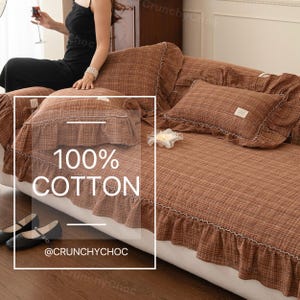 May include: Brown plaid sofa cover with frilled edges and matching cushions. The cover is made of 100% cotton and has a white trim. The text "100% COTTON" is displayed on the cover. The brand name "@CRUNCHYCHOC" is also visible.