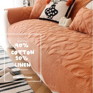 May include: A terracotta-colored sofa cover featuring a quilted swirl pattern and a scalloped edge. The fabric is labeled as 90% cotton and 10% linen. A white decorative pillow with a black vase graphic and a small white beaded bag sit on the sofa.