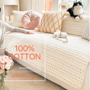 以下が含まれることがあります： 「100% COTTON」の文字が入った、クリーム色のキルティングソファカバー。丸いピンクのクッションや、オレンジと白の模様のクッションなど、様々な形と色の装飾用クッション。黒と白のぬいぐるみ猫がソファに座っています。