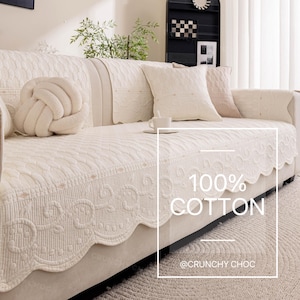 May include: Cream-colored sofa cover with a scalloped edge and embroidered details. The cover is made of 100% cotton and includes matching throw pillows and a decorative knot pillow. The text "100% COTTON" is visible on the cover.