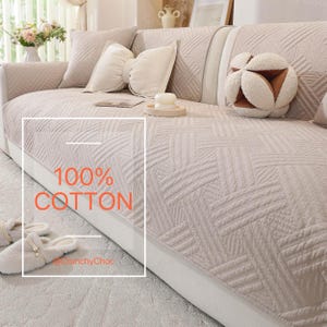 May include: Beige sofa cover with a geometric pattern, accompanied by matching cushions and a decorative ball. The cover is labelled "100% COTTON". The overall aesthetic is soft and inviting, suitable for living room decor.