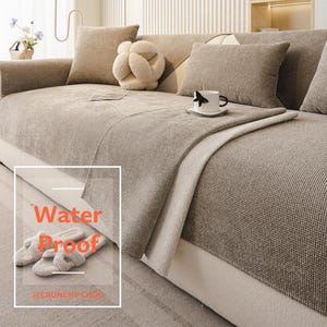 May include: A beige textured sofa cover with a waffle-knit pattern draped over a couch. The cover features a rectangular overlay with the text Water Proof and @CRUNCHY CHOC. Includes matching beige throw pillows and a white mug on the seat.