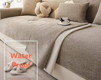 Pode incluir: Uma capa de sofá bege texturizada com padrão de tecido waffle sobre um sofá. A capa apresenta uma sobreposição retangular com o texto Water Proof e @CRUNCHY CHOC. Inclui almofadas bege a condizer e uma caneca branca no assento.