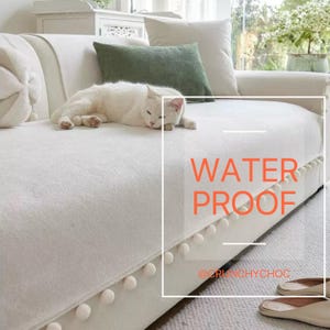 Pode incluir: Um sofá branco com um gato branco descansando sobre ele. O sofá tem uma superfície texturizada e é adornado com uma fileira de pompons brancos na borda inferior. O texto "WATER PROOF" é visível.