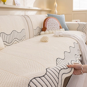 May include: Cream-colored quilted sofa cover with a wavy black and white border design. The cover features a chevron pattern and is adorned with decorative pillows, including a rainbow-shaped pillow and a blue square pillow.